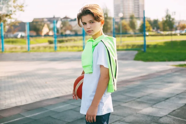 Seorang remaja cantik memegang bola basket di tangannya dan melihat ke kamera. Potret seorang anak laki-laki dengan bola di tangannya. Olahraga, pendidikan, kesenangan — Stok Foto