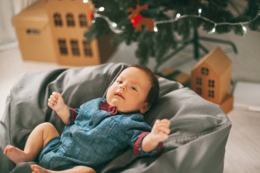 Küçük bir çocuk Noel ağacının yanındaki bir puf üzerinde uzanıyor ve gülümsüyor. Çocuk tatilden memnun kaldı. Tatil, etkinlik.