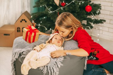 Ren geyiği kostümü giymiş küçük bir çocuk bir Noel ağacının yanındaki battaniyenin üzerinde yatıyor ve hediyeler alıyor. Kız kardeşi de yanında oturuyor ve onu öpüyor. Tatil, etkinlik, dekorasyon. Noel ve Yeni Yıl
