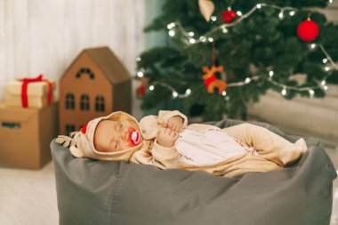 Ren geyiği kostümlü çekici bir bebek Noel ve yeni yıl için dekore edilmiş bir odada puf üzerinde uyur. Bir çocuk Noel arifesinde bir Noel ağacının yanında uyur. Tatil, olay, metin için yer