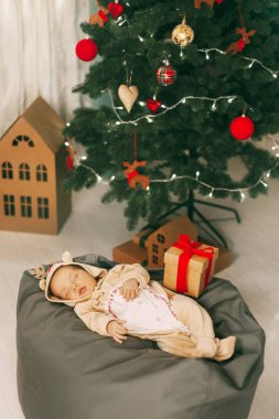 Ren geyiği kostümlü çekici bir bebek Noel ve yeni yıl için dekore edilmiş bir odada puf üzerinde uyur. Bir çocuk Noel arifesinde bir Noel ağacının yanında uyur. Tatil, olay, metin için yer