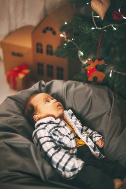 Bebek bir puf üzerinde yatıyor ve evdeki Noel ağacının süslerine bakıyor. Noel 'de ve yeni yıl tatillerinde küçük tatlı bir çocuk. Tatil, etkinlik, Noel, yeni yıl, yer