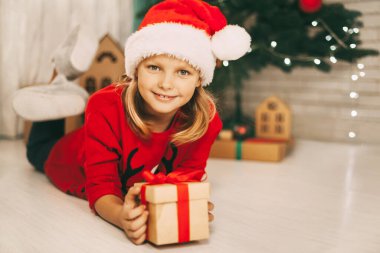 Noel Baba şapkalı güzel sarışın bir kızın portresi. Elinde bir hediyeyle yerde yatıyor. Süslü bir Noel ağacının arka planında. Noel ve Yeni Yıl konsepti. Tatil, hediye.