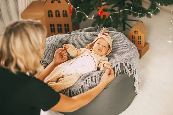 A beautiful baby in a deer costume lies on a knitted blanket and holds his mothers hands. Child and mother on the background of a decorated Christmas tree. New year holiday. Christmas and New year — Stock Photo, Image