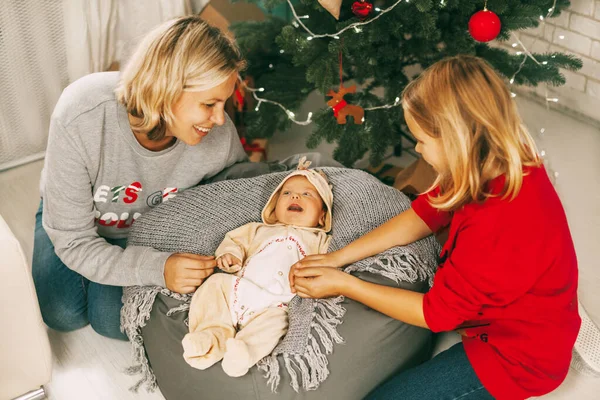 A cheerful baby lies next to the Christmas tree and smiles at his mother and sister. A mother and a girl play with a baby in a room decorated for Christmas. Holiday, New year, Christmas — Stock Photo, Image