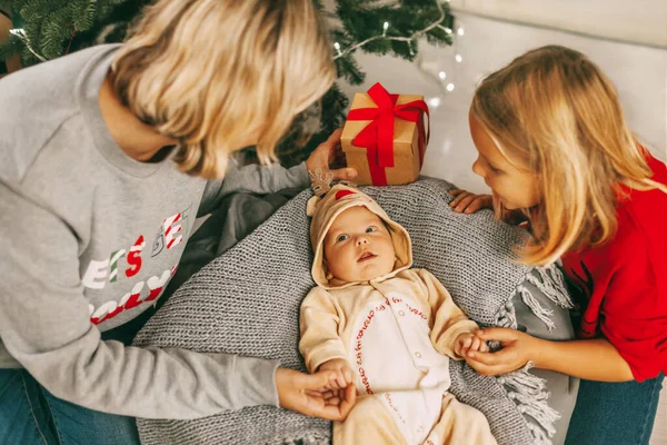 A cheerful baby lies next to the Christmas tree and smiles at his mother and sister. A mother and a girl play with a baby in a room decorated for Christmas. Holiday, New year, Christmas — Stock Photo, Image