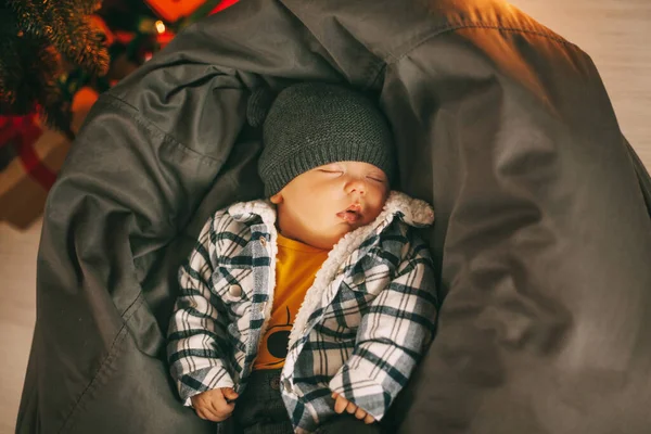 En søt nyfødt baby i rutete jakke og en strikket hatt sover på en pouf ved siden av et juletre. Nye år. Plass til tekst – stockfoto