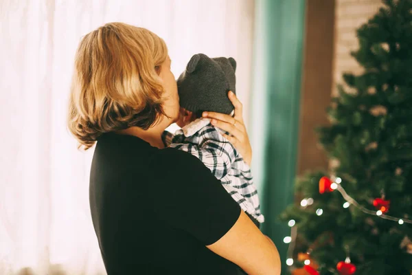 En mor håller sin unge son i famnen och kysser honom. Baby med mamma i ett festligt dekorerat rum med en julgran. Semester, evenemang, plats för text. Jul och nyårskoncept, bakifrån — Stockfoto