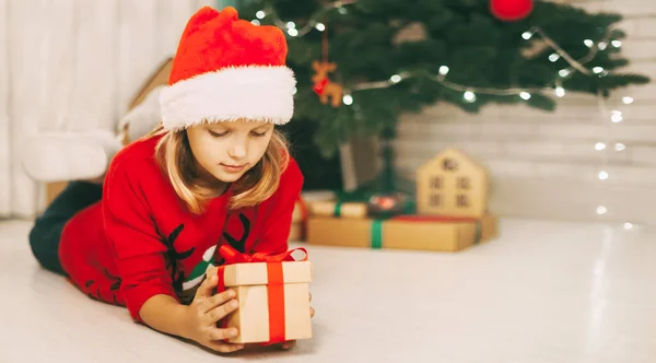 Una chica rubia yace en el suelo junto a un árbol de Navidad decorado y sostiene un regalo atado con una cinta roja. La chica desenvuelve el regalo. Navidad y año nuevo concepto —  Fotos de Stock