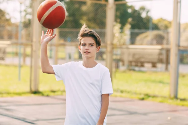 Um jovem jogador de basquete adolescente em uma camiseta branca fica na quadra de basquete, girando uma bola de basquete em seus dedos. O conceito de esporte e um estilo de vida saudável — Fotografia de Stock
