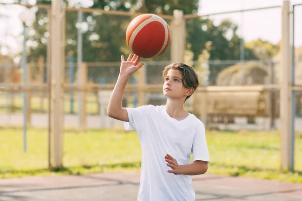 Seorang pemuda pemain basket dengan kaos putih berdiri di lapangan basket, memutar-mutar bola basket di jari-jarinya. Konsep olahraga dan gaya hidup sehat — Stok Foto