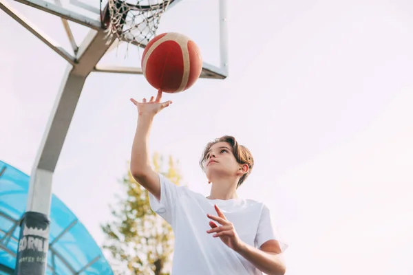 Seorang pemuda pemain basket dengan kaos putih berdiri di lapangan basket, memutar-mutar bola basket di jari-jarinya. Konsep olahraga dan gaya hidup sehat — Stok Foto