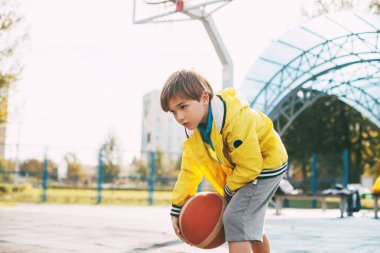 Sarı spor ceketli şirin bir çocuk basketbol topu fırlatıyor. Bir çocuk basketbol oynuyor. Spor, eğitim, yaşam tarzı