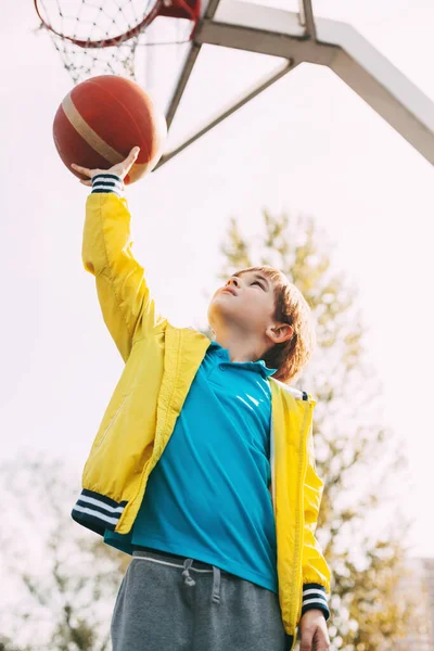 Portrait d'un mignon garçon joueur de basket-ball debout avec une balle dans les mains à côté du panier de basket-ball. Le concept du sport et un mode de vie sain — Photo