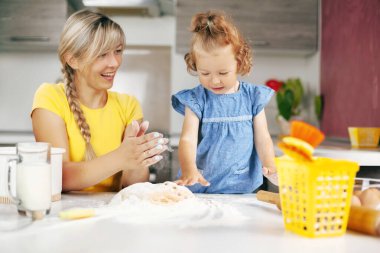 Genç ve güzel bir kadın ve küçük kızı mutfakta kurabiye hamuru yoğuruyorlar. Anne ve kızı gülümser ve birlikte yemek pişirirler. Aile eğlencesi, birlikte eğlence.