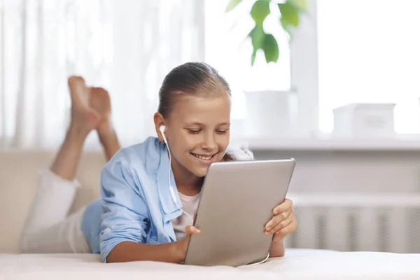 Chica adolescente rubia acostada en el sofá juega un juego en la tableta y se comunica a través de un enlace de vídeo con amigos. Tecnologías digitales —  Fotos de Stock