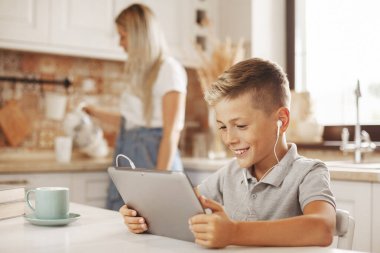 Çocuk ödevini mutfakta, internet tabletinde öğretmeniyle konuşarak yapıyor. Anne yemek pişirir ve oğluna yardım eder. Uzaktan öğrenme, çevrimiçi eğitim, evde eğitim kavramı