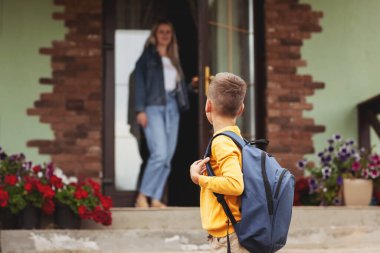 Anne, evinin yanındaki kapıda duruyor, oğluna okula kadar eşlik ediyor, okula geri dönüyor..