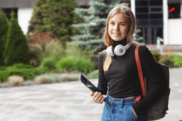 Una hermosa estudiante femenina de moda en auriculares se encuentra en la ciudad con una tableta y una mochila y sonrisas. Retrato — Foto de Stock