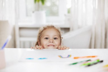 Küçük bir kız masanın altına saklanır ve altından dışarı bakar. Kız ev ödevleri arasında arada oynuyor. Ev ödevi, ev eğitimi, online öğrenim.