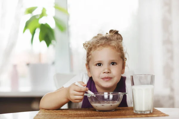 Ragazza affascinante mangia cereali con latte per la prima colazione e. Sul tavolo ci sono fiocchi di cioccolato, un bicchiere di latte. Colazione sana, prendersi cura dei bambini. Spazio per testo — Foto Stock