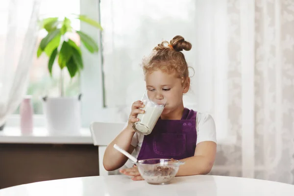 Carina bambina mangia cereali con latte per colazione. La ragazza beve latte da un bicchiere di vetro. Colazione sana, prendersi cura dei bambini. Spazio per testo — Foto Stock