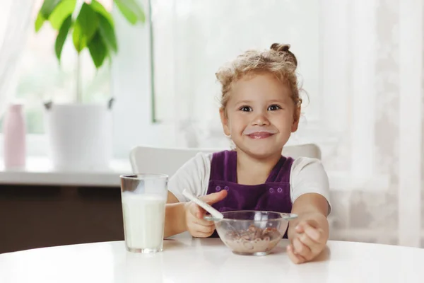 Ragazza affascinante mangia cereali con latte per la prima colazione e. Sul tavolo ci sono fiocchi di cioccolato, un bicchiere di latte. Colazione sana, prendersi cura dei bambini. Spazio per testo — Foto Stock