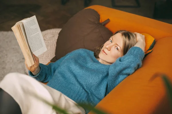 Una hermosa chica se está relajando en el sofá de la sala de estar, leyendo su novela favorita. Linda y pensativa chica está tumbada en el sofá y leyendo un libro. Trabajo en línea, freelance, distancia social —  Fotos de Stock