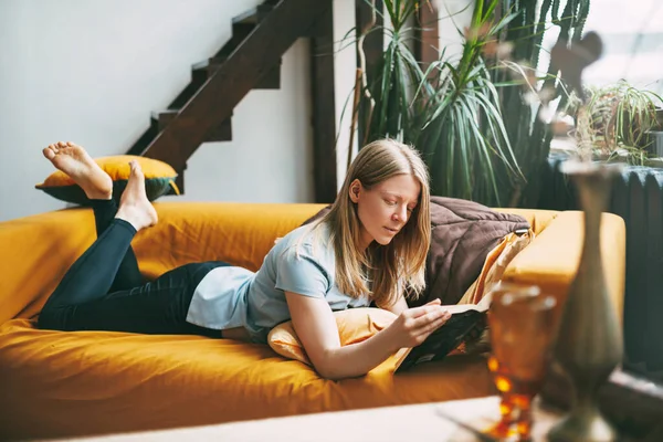Uma jovem está deitada no sofá e lendo um livro favorito. A menina está descansando, dedica seu tempo livre para ler. — Fotografia de Stock