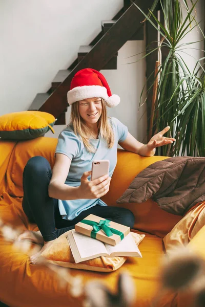 Una chica sonriente en un sombrero de Navidad se sienta en el sofá y se comunica a través de un enlace de vídeo con amigos. Chica felicita a amigos y familiares en vacaciones —  Fotos de Stock