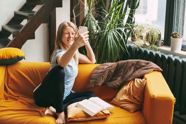 Uma jovem está sentada no sofá e tomando uma selfie em seu telefone, um livro aberto está deitado ao lado dela — Fotografia de Stock