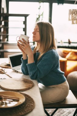 Genç bir kadın sabahları kahvesinden zevk alır ve dizüstü bilgisayarında çalışır. Evde çalışmak, online iş, serbest çalışmak. Yan görünüm