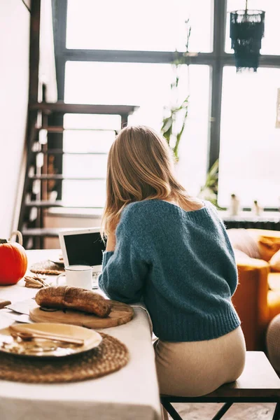 A blonde girl works in the morning at a laptop and drinks coffee. Work online, freelance, social distance. Rear view — Stock Photo, Image