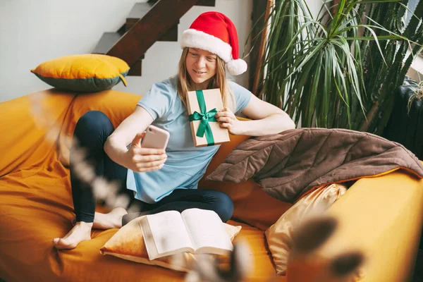 A smiling girl in a Christmas hat sits on the sofa and communicates via video link with friends and shows them her gift.