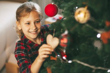 Yeni yıl kıyafetleri içinde küçük mutlu bir sarışın, Noel ağacını süslüyor ve gülümsüyor. Yılbaşı ve evde yeni yıl tatili konsepti