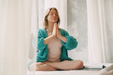 Genç bir kız, yatak odasındaki yatakta Lotus pozisyonunda otururken meditasyon yapıyor. Kız sabahları evde stres önleyici yoga yapıyor..