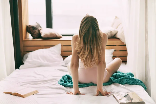 A beautiful girl sits on the bed in the early morning and stretches after waking up, next to an open book. Rear view.