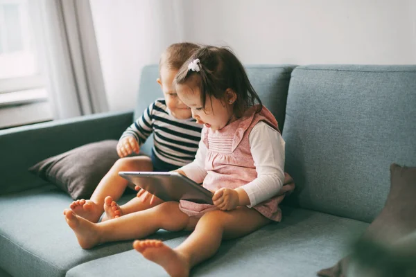 Dois bebês adoráveis estão sentados no sofá e brincando em um tablet. Irmão e irmã passam o tempo brincando juntos — Fotografia de Stock