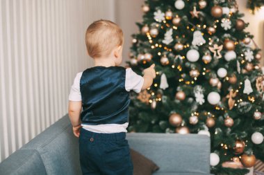 Takım elbiseli küçük bir çocuk kanepede duruyor ve Noel ağacında asılı olan oyuncakları işaret ediyor. Dikiz aynası. Tatil, etkinlik, Noel ve Yeni Yıl