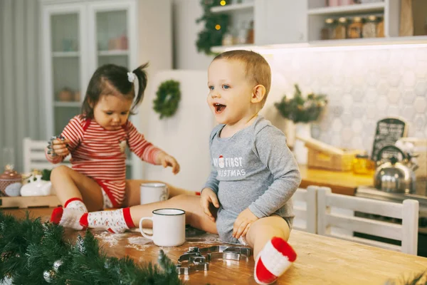 Funny kids sit on the kitchen table and play with cookie cutters. Children help bake cookies for Christmas and New year — Stock Photo, Image