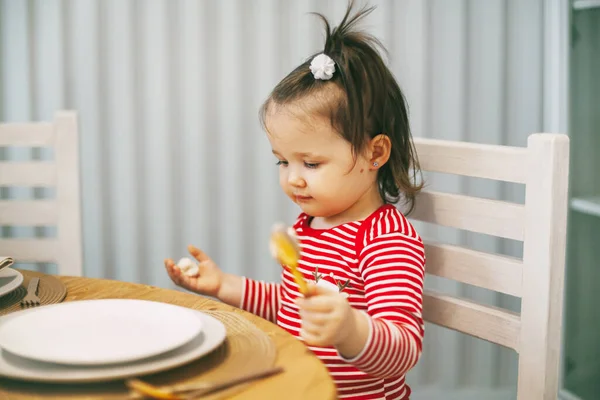 Affascinante bambina seduta a tavola con un cucchiaio in mano in attesa della cena di Natale. — Foto Stock