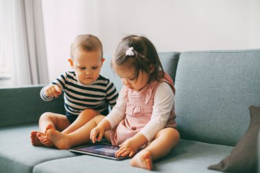 İki sevimli bebek kanepede oturuyor ve tablette oynuyorlar. Erkek ve kız kardeş birlikte vakit geçiriyorlar.