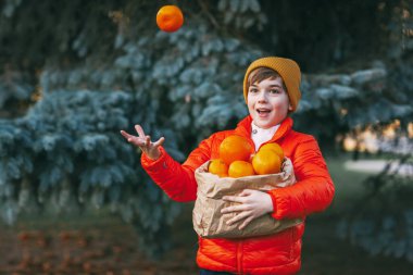 Parlak turuncu ceketli ve sarı şapkalı bir çocuk elinde büyük bir torba portakal tutuyor ve bir Noel ağacının arka planına gülüyor ve portakal fırlatıyor. Noel ve Yeni Yıl için Hazırlık