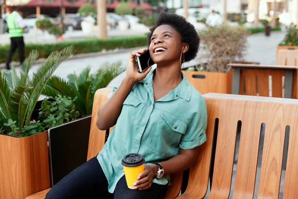 Jovem africana sentada em um banco, bebendo café e falando ao telefone — Fotografia de Stock