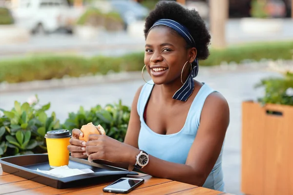 Mulher africana bonito almoçar ao ar livre no café da cidade. Uma mulher afro-americana sorri enquanto se senta em uma mesa em um café da cidade, segurando um hambúrguer, ao lado de um copo de café — Fotografia de Stock