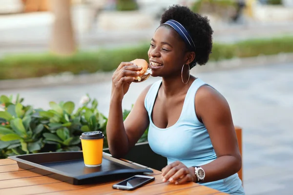 Mulher africana bonito almoçar ao ar livre no café da cidade. Uma mulher afro-americana sorri enquanto se senta em uma mesa em um café da cidade, segurando um hambúrguer, ao lado de um copo de café — Fotografia de Stock