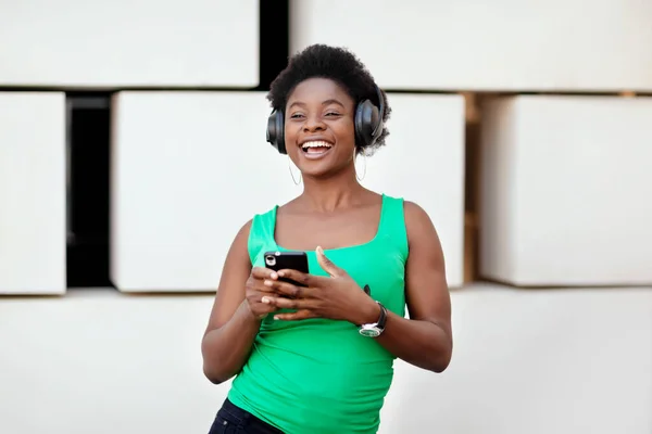 Menina africana feliz ouve música com fones de ouvido sem fio em seu telefone celular e danças. A menina está gostando da música. — Fotografia de Stock