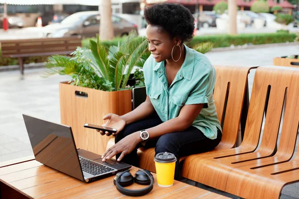 通りのカフェに座ってコンピュータで仕事をしているアフリカ人の少女の笑顔。少女はノートパソコンと電話でデータをチェックします。オンラインで仕事、ビジネスオンライン — ストック写真