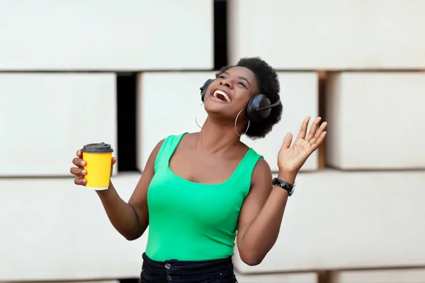 Mulher africana feliz segurando um café em uma Copa descartável, ouvindo a melodia agradável nos fones de ouvido e dança. Está na hora da música. Espaço para texto — Fotografia de Stock