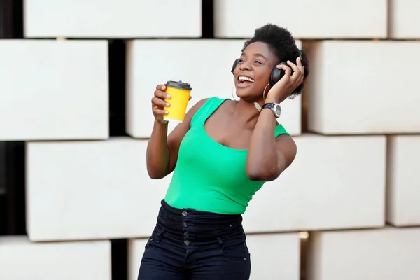 Mulher africana sorridente segura café em uma xícara descartável, ouve uma melodia agradável em fones de ouvido e danças — Fotografia de Stock